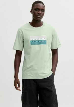 Mężczyzna ubrany w jasnozieloną koszulkę Jack & Jones i czarne szorty typu cargo, stoi z jedną ręką w kieszeni na jednolitym tle.