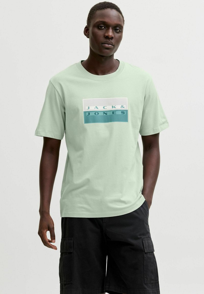 Homme portant un t-shirt vert clair Jack & Jones et un short cargo noir, debout avec une main dans la poche devant un fond uni.