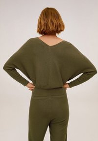 Personne aux cheveux courts châtain clair, debout avec les mains sur les hanches, portant un haut à manches longues vert olive et un pantalon assorti, tournée dos à l'appareil photo.