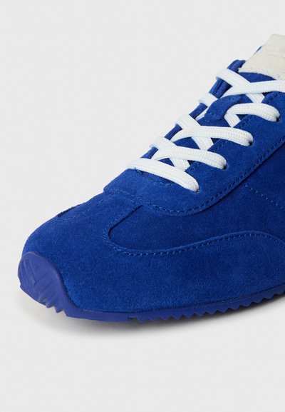 Modrá semišová teniska s bielymi šnúrkami, vybavená zaoblenou špičkou, textúrovaným zvrškom a gumovou podrážkou so vzorom cik-cak pre lepšiu priľnavosť.