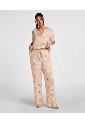 Vrouw staat blootsvoets en draagt een pastelroze pyjamaset met fruitpatronen, een kort mouwen knoopshirt en losse broek met trekkoord.