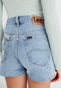 Shorts en denim bleu clair avec bords effilochés, présentant une broderie florale sur la poche arrière. Étiquette en cuir marron "Lee" au niveau de la taille.