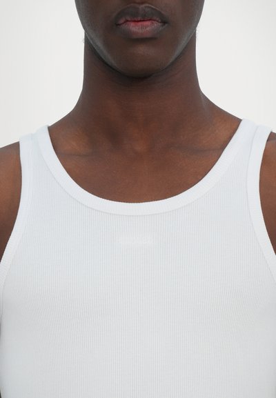 Débardeur blanc à côtes avec un col rond. Présente un logo discret d'une teinte plus claire sur le devant. Le tissu semble lisse et léger.