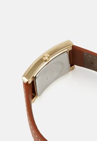 Montre rectangulaire dorée avec un dos en métal argenté, un bracelet en cuir marron et une couronne ronde sur le côté.