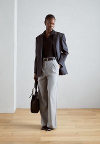 Pantalon large gris avec plis, associé à un polo noir et une veste à rayures. Accentué par une ceinture et tenant un sac à main noir.
