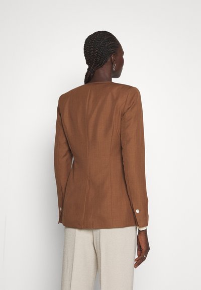 Γυναίκα με πλεξούδες, που φοράει ένα εφαρμοστό καφέ σακάκι και ανοιχτό μπεζ παντελόνι, που στέκεται μπροστά σε έναν απλό λευκό τοίχο, γυρτώντας προς τα πίσω.