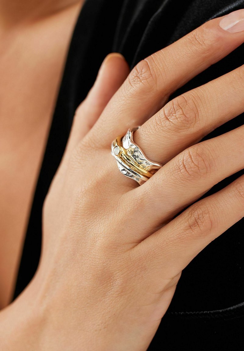 Anillo de declaración en plata y oro con diseño ondulado; presenta superficies pulidas y texturizadas, colocado en un dedo con un tono de piel natural.
