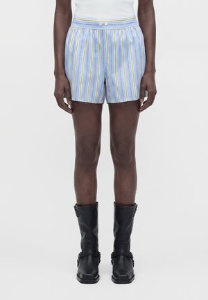 Personne portant un short rayé bleu clair avec un détail bouton, un haut blanc et des bottes en cuir noires mi-mollet, debout devant un fond uni.