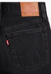 Černé džínové kalhoty z denimu s koženou nášivkou na pasu s logem „Levi Strauss & Co.“, výrazným prošitím zadních kapes a červenou štítkovou ozdobou.