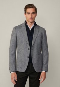 Blazer gris texturizado con dos botones, solapas de muesca y bolsillos frontales. Llevado sobre un suéter azul marino y una camisa blanca con cuello con cremallera.