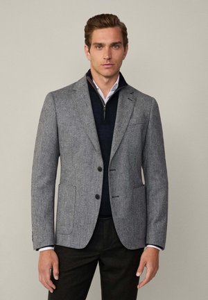 Grauer Blazer mit Textur, zwei Knöpfen, Revers und frontalen Taschen. Getragen über einem navy-blauen Pullover und einem weißen Hemd mit einem Reißverschlusskragen.