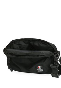 Černá látková ramenní taška se zipovým uzávěrem, nastavitelným popruhem a vnitřními přihrádkami. Na přední straně je logo.