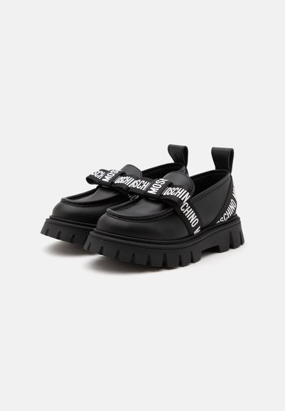 Μαύρα παπούτσια slip-on με χοντρή σόλα και φαρδιές λωρίδες, με λευκή επιγραφή Moschino στο πάνω μέρος και πίσω.