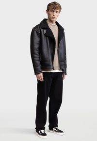 Giacca aviator in pelle nera con collo in shearling, indossata sopra un maglione beige a maglia, abbinata a pantaloni neri a vestibilità ampia e sneakers nere.