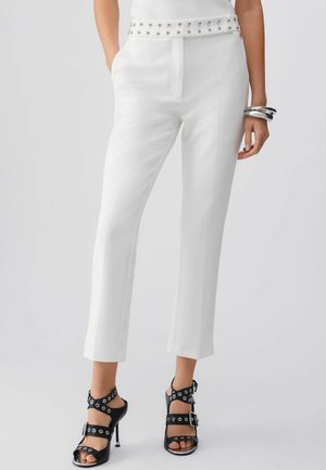 Białe dopasowane spodnie z paskiem z ćwiekami noszone z czarnymi sandałami na wysokim obcasie z licznymi klamrami i oczkami, srebrne bransoletki na nadgarstku.