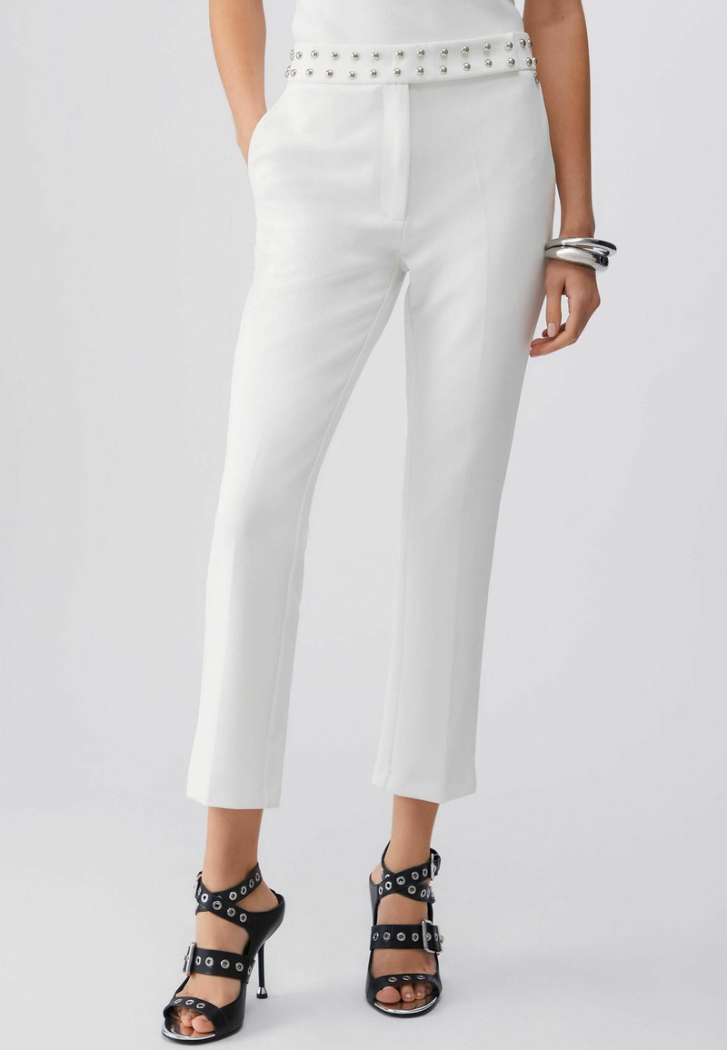 Valged kohandatud püksid koos naelutatud vöökohaga, kantud mustade kõrgete kontsadega sandaalidega, millel on mitu pandlat ja silmaauku, ning hõbedased käevõrud randmel.