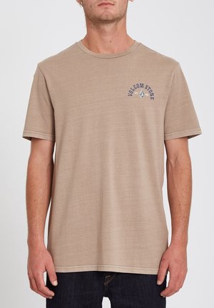 Mężczyzna ubrany w gładki beżowy t-shirt z krótkim rękawem i małym granatowym logo Volcom Stone na lewej piersi, zestawiony z ciemnymi dżinsami.