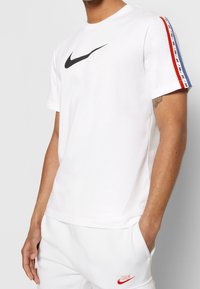 Bílý bavlněný tričko s černým logem Nike, červenými a modrými akcentními pruhy na rukávech, kulatým výstřihem a krátkými rukávy.