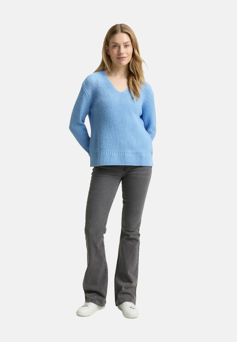 Frau steht mit Händen hinter dem Rücken, trägt einen hellblauen Strickpullover, graue Bootcut-Hosen und weiße Sneakers vor einem weißen Hintergrund.