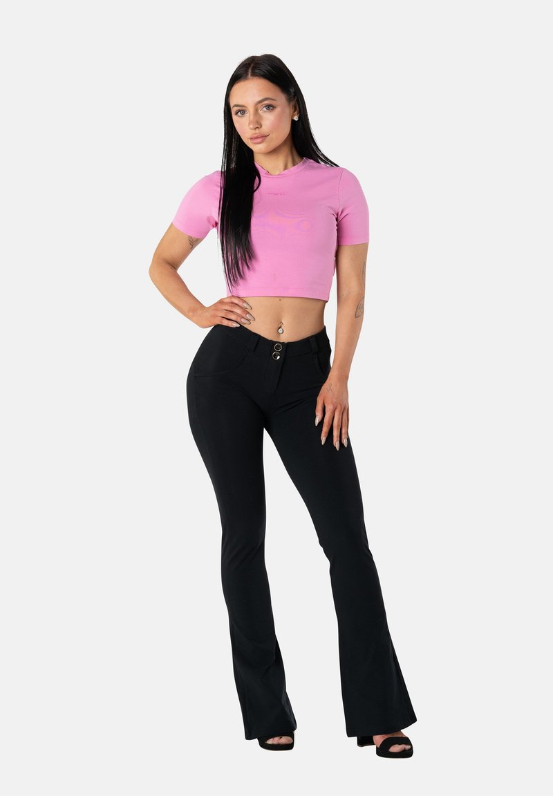 Giovane donna con lunghi capelli scuri che indossa un top corto rosa, pantaloni neri aderenti e svasati e tacchi neri aperti, in piedi con una mano sul fianco.