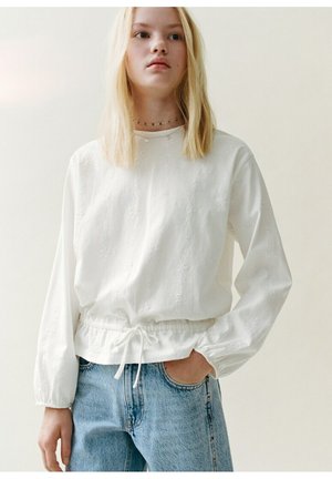 Mladá žena s dlhými blond vlasmi má na sebe bielu blúzku s dlhým rukávom so zaväzovaním v páse a svetlomodré džínsové nohavice, stojí s jednou rukou v vrecku.