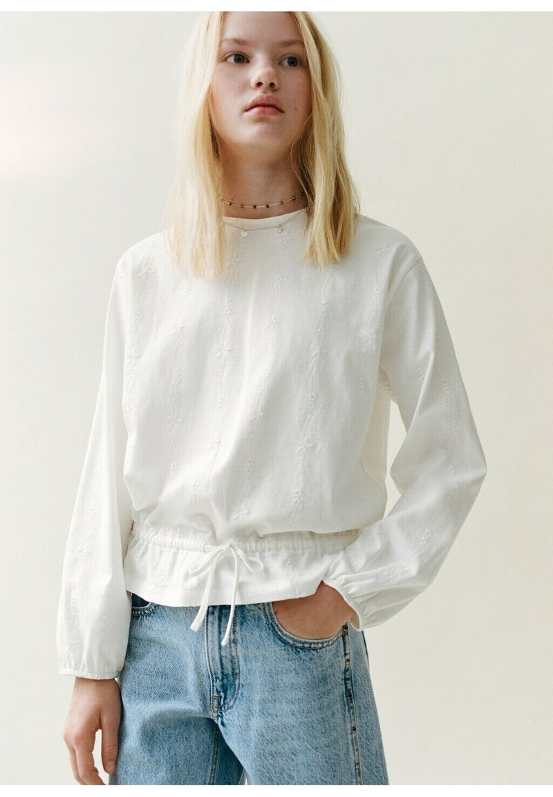 Mladá žena s dlhými blond vlasmi má na sebe bielu blúzku s dlhým rukávom so zaväzovaním v páse a svetlomodré džínsové nohavice, stojí s jednou rukou v vrecku.