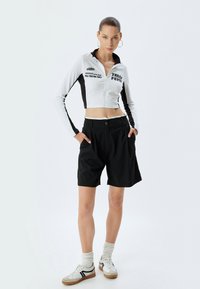 Giacca cropped con zip bianca e nera con testo, abbinata a pantaloni corti neri. Il modello indossa calze bianche e sneakers in stile retrò con dettagli.