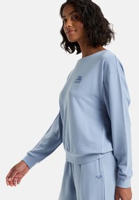 Světle modrý svetr z měkké tkaniny s volným střihem. Disponuje kulatým výstřihem a vyšitým logem na prsou. Obsahuje žebrované manžety.