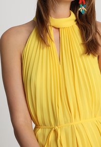 Robe jaune plissée à col halter avec une ouverture en forme de trou de serrure. Fabriquée en tissu léger, elle présente une taille à nouer et une texture lisse.