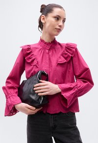 Φούξια μπλούζα με βολάν στον γιακά και τα μανίκια, διακοσμητική δαντέλα και κλείσιμο με κουμπιά. Κρατάει μαύρη δερμάτινη τσάντα. Σκούρο τζιν.