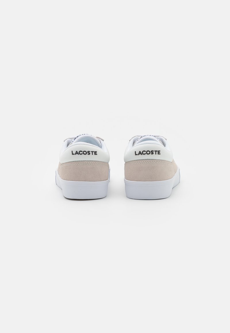 Une paire de baskets Lacoste beige et blanche vue de l'arrière, avec le nom de la marque sur le col du talon, sur fond blanc.