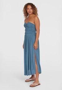 Himmlisch hellblaues trägerloses Maxikleid mit gerüschtem Oberteil und frillenden Akzenten, das seitliche Schlitze und eine fließende Textur aufweist. Getragen mit flachen Sandalen.
