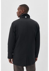 Cappotto nero di lunghezza media con colletto alto, caratterizzato da una texture liscia e bottoni a scatto laterali. Etichetta con logo posizionata sulla parte posteriore del collo.