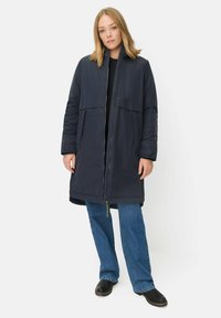 Kvinde iført en lang, marineblå, polstret frakke, blå jeans og sorte sko, der står foran en ensfarvet lys baggrund.