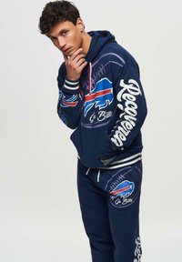 Mörkblå hoodieset och byxor med lagets logotyp, grafiska mönster och randiga manschetter. Materialet verkar mjukt med en avslappnad passform. Röda detaljer finns.