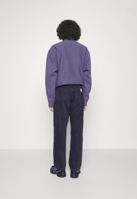 Henkilö, jolla on kiharaiset hiukset, pukeutuneena violettiin pitkähihaiseen paitaan, mustiin farkkuihin ja mustiin lenkkareihin, seisoo kasvot kohti yksinkertaista valkoista seinää.