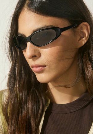 Jauna moteris su ilgais tamsiais plaukais, dėvinti siaurus juodus kačių akies formos saulės akinius ir tamsų ribotu tekstūru viršutinį drabužį su šviesia striuke.