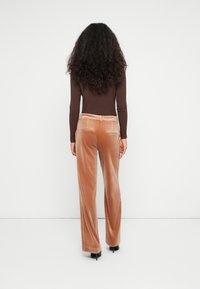 Mujer con cabello rizado y largo de pie frente a una pared blanca, vestida con una blusa marrón de canalé, pantalones de terciopelo beige de pierna ancha y zapatos de tacón negros.