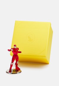 Translucent red action figure on a circular clear base, next to a textured yellow box with a raised logo.