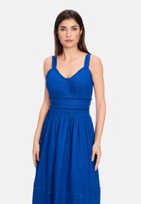 Blaues Kleid aus leichtem Stoff, mit einem figurbetonten Oberteil mit vertikalen Falten und einem ausgestellten Rock. Dünne Träger und geraffte Kanten.