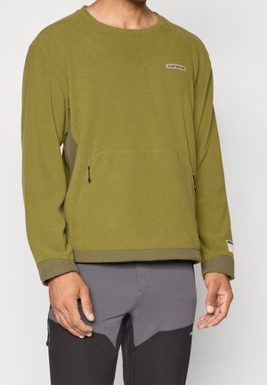 Mann trägt olivgrünen Langarm-Pullover mit vorderen Reißverschlusstaschen und graue und schwarze Sporthose, steht in neutraler Pose.