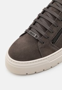 Gros plan d'une basket en daim marron avec une semelle blanche épaisse, un embout rond, des lacets assortis, une étiquette de lacet de marque et une fermeture éclair latérale noire.