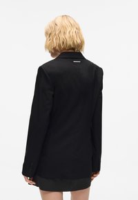 Melnā žakete ar pielāgotu piegriezumu, ar vienu venti aizmugurē un pogu akcentiem uz piedurknēm. Gluda auduma tekstūra ar vienkāršu dizainu.