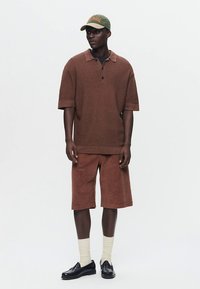 Hnedé textúrované polo tričko s krátkym rukávom a golierom, skombinované so zodpovedajúcimi krátkymi nohavicami z manžestru. Nosené so zelenou čiapkou a čiernymi topánkami.