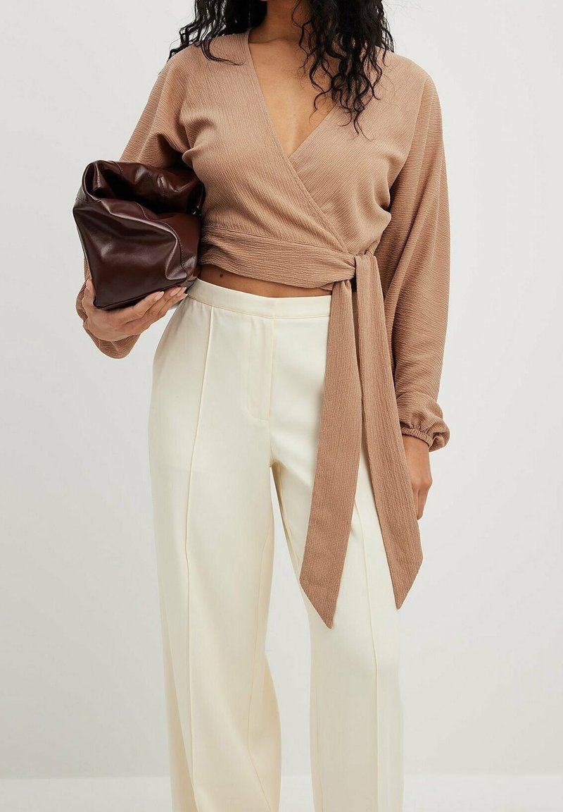 Beige ribbet indpakningsbluse med lange ærmer og en snøring, parret med cremefarvede vide bukser. Modellen holder en brun, krøllet lædertaske.