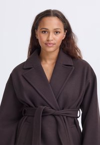 Giovane donna con lunghi capelli che indossa un cappotto scuro con cintura, in piedi contro uno sfondo chiaro e semplice, che guarda verso la telecamera.