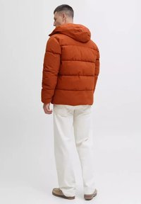 Puffer jacket in orange, featuring a hood and quilted sections. Paired with white pants and beige shoes, viewed from the back.
