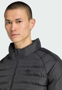 Detailný záber muža s krátkymi vlasmi, ktorý má na sebe čiernu pufovanú bundu s vysokým golierom a logom Adidas na hrudi.