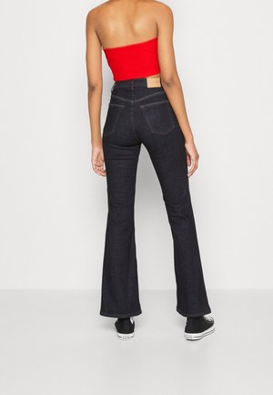 Dunkelblaue, ausgestellte Jeans mit taillierter Passform, ausgestattet mit einem Lederpatch auf der Rückseite und Kontrastnähten. Kombiniert mit einem roten, cropped Top und schwarzen Sneakers.