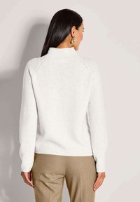 Frau mit langen dunklen Haaren, die goldene Creolenohrringe, einen weiß gesprenkelten Strickpullover und beige strukturierte Hosen trägt, von hinten gezeigt.